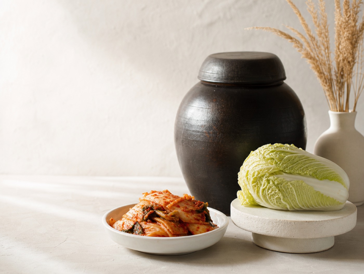 An onggi crock, a whole napa cabbage and a dish of fresh kimchi on a pale surface